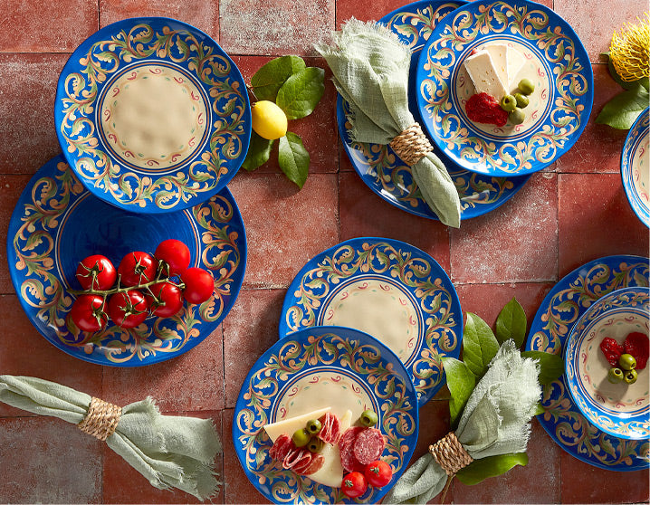 Top-down view of blue and gold ornate plates arranged with cheese, cherry tomatoes and greens on a red tiled table
