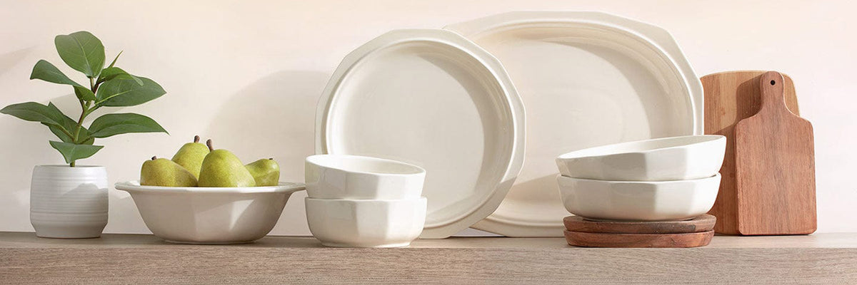 Heritage white ceramic dinnerware with stacked bowls large plates cutting boards a bowl of pears and a potted plant.