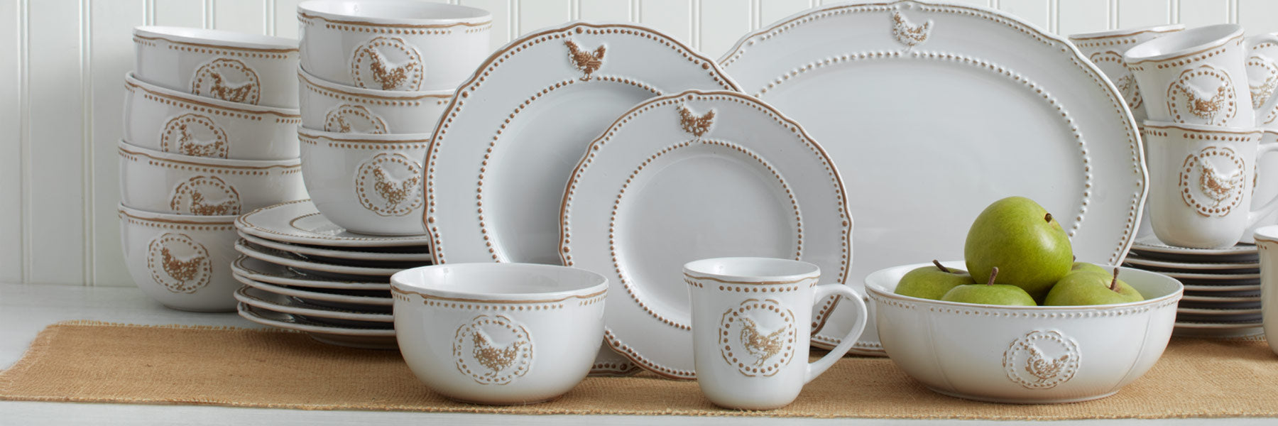 Farmhouse Collection white ceramic dinnerware with beaded edges and rooster motif, stacked bowls and plates with a mug and fruit bowl on a burlap runner.