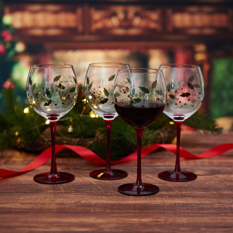 Four Winterberry hand painted wine goblets with holly leaf pattern on a wooden table, one filled with red wine.