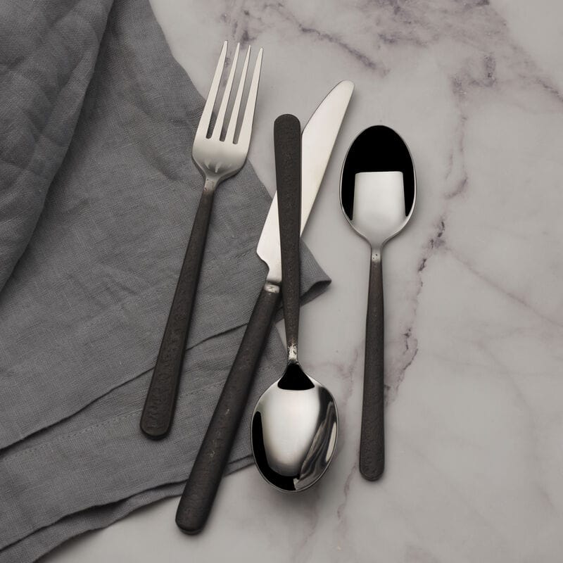 Calyx Black Forged 20 piece flatware set for four displayed on marble with a grey napkin and dark textured handles