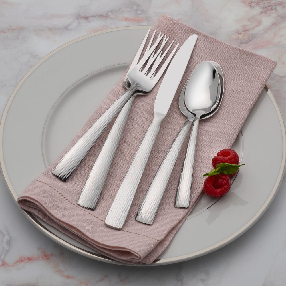 Hammered stainless steel flatware arranged on a blush napkin atop a pale plate with two raspberries.