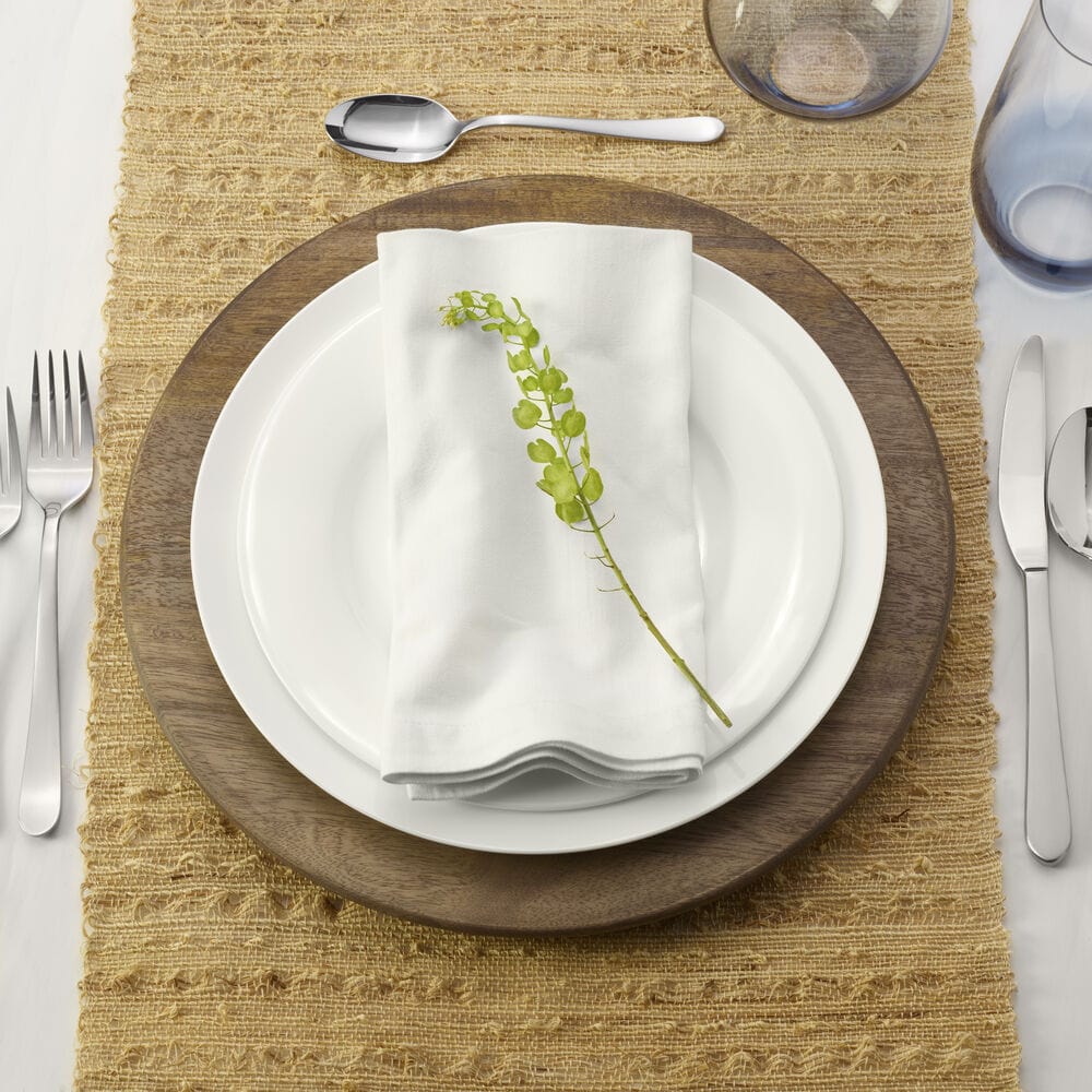 Top view of a dining place setting with white plates on a round mango wood charger and a folded napkin with greenery