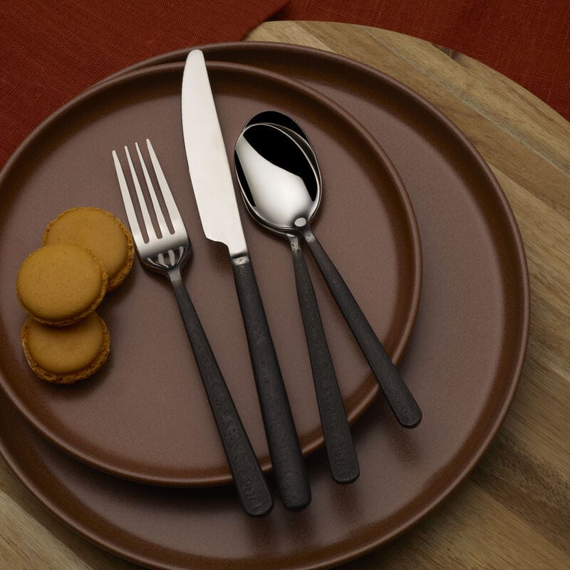 Calyx Black Forged 20 piece flatware set arranged on a brown plate with knife, fork, and spoon beside three macarons