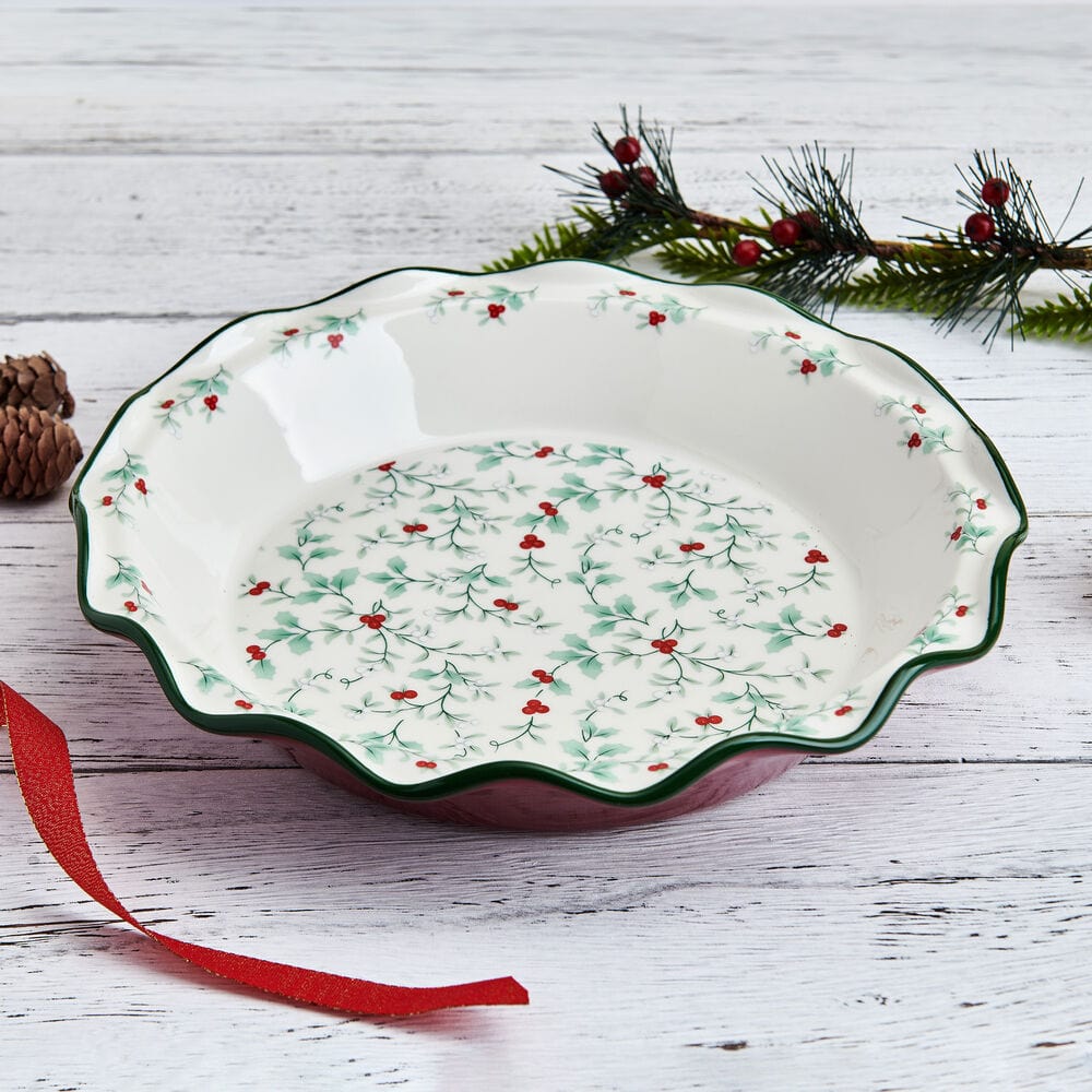 Winterberry Pie Plate with holly motif and green rim on a white wooden surface with pinecones and red ribbon.