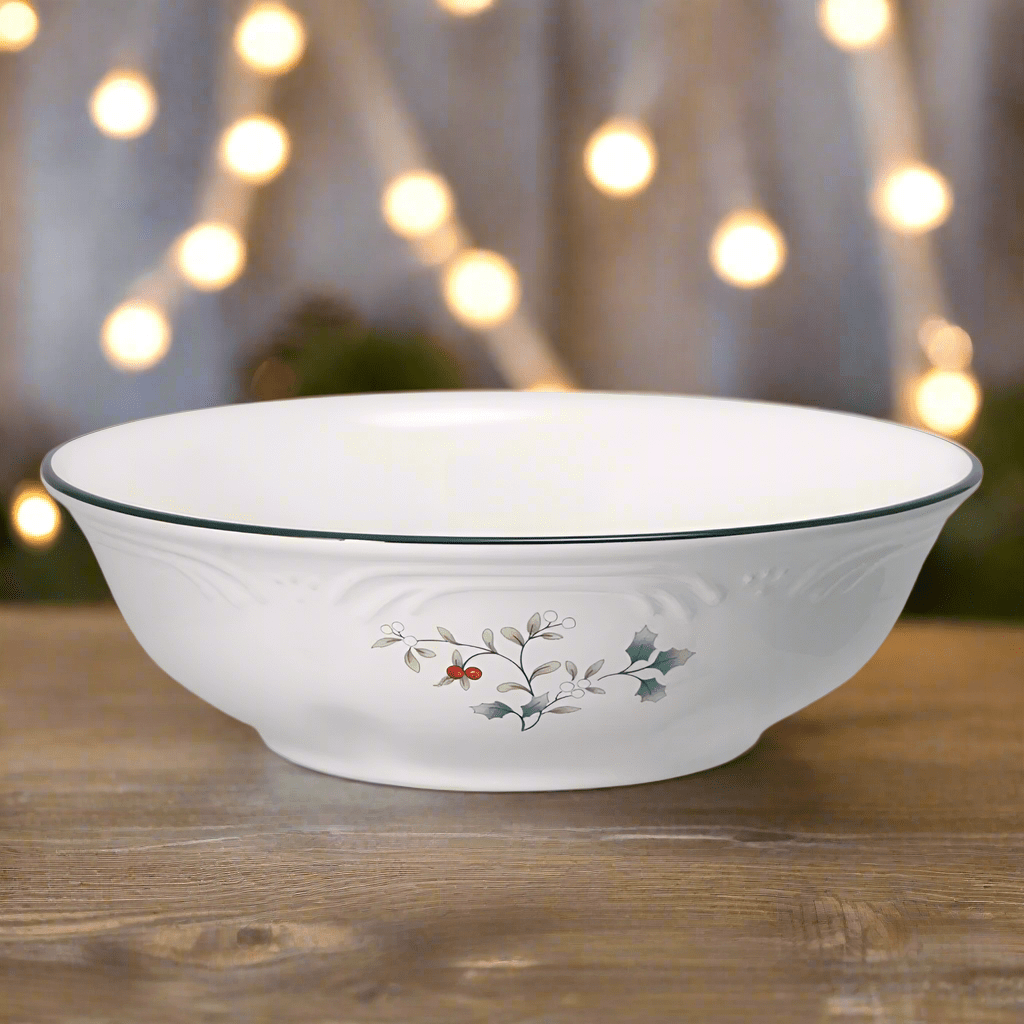 Winterberry vegetable serve bowl, white ceramic with holly berry motif and dark rim on a wooden table with warm bokeh background.