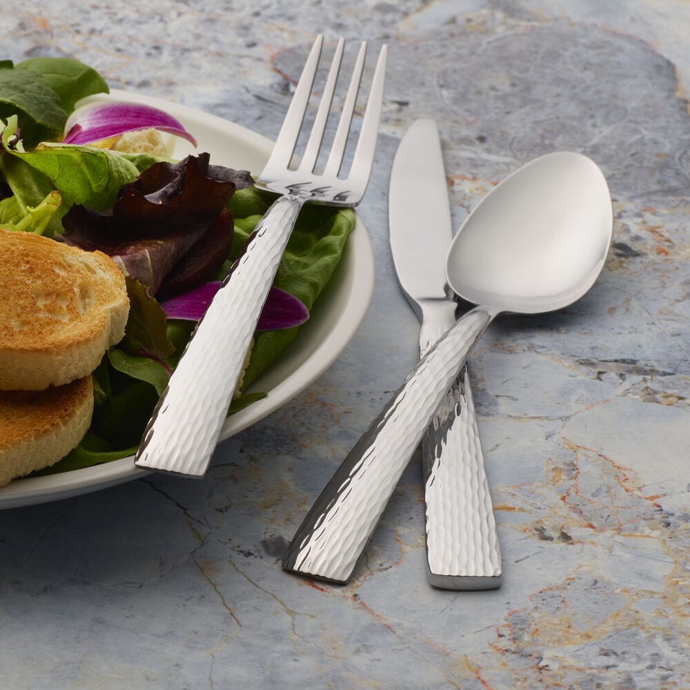 Plate with greens and bread beside hammered Elliott flatware laid across a marble surface.