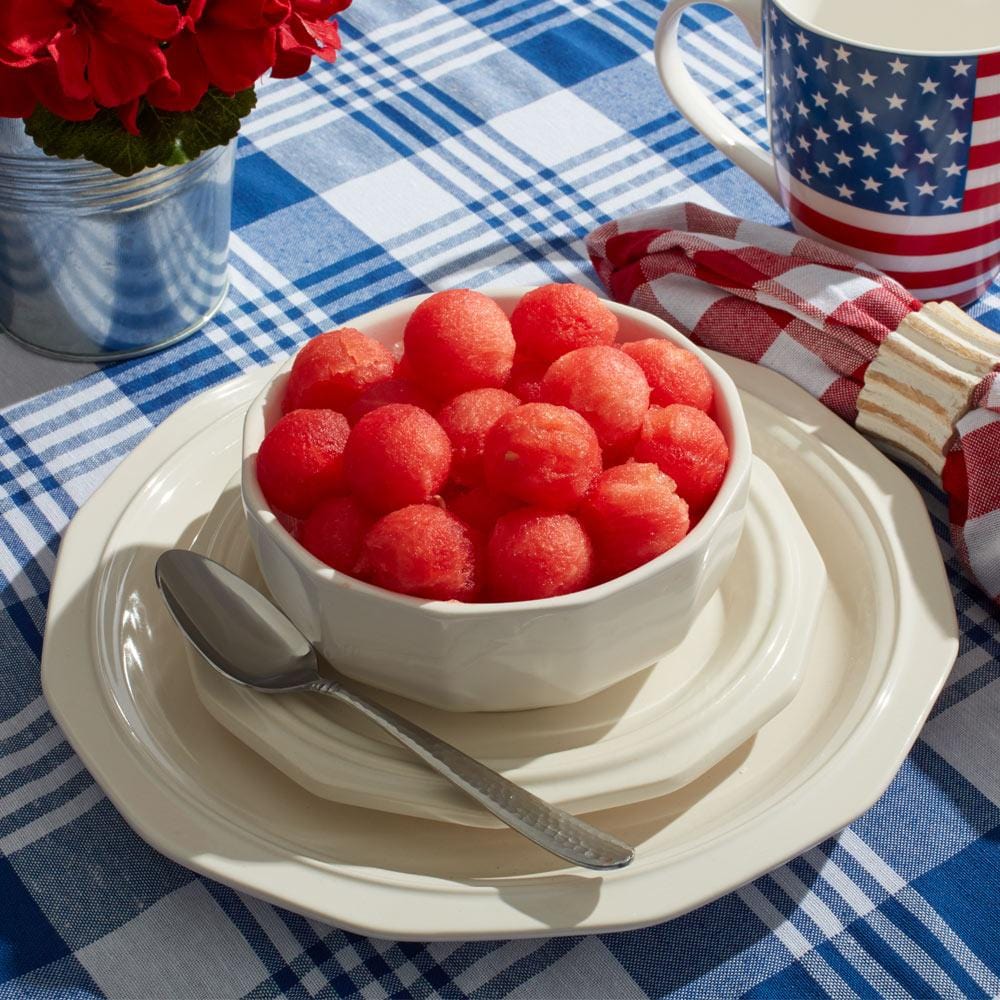 Heritage bowl filled with red melon balls on a plate beside a patriotic mug and blue plaid tablecloth.