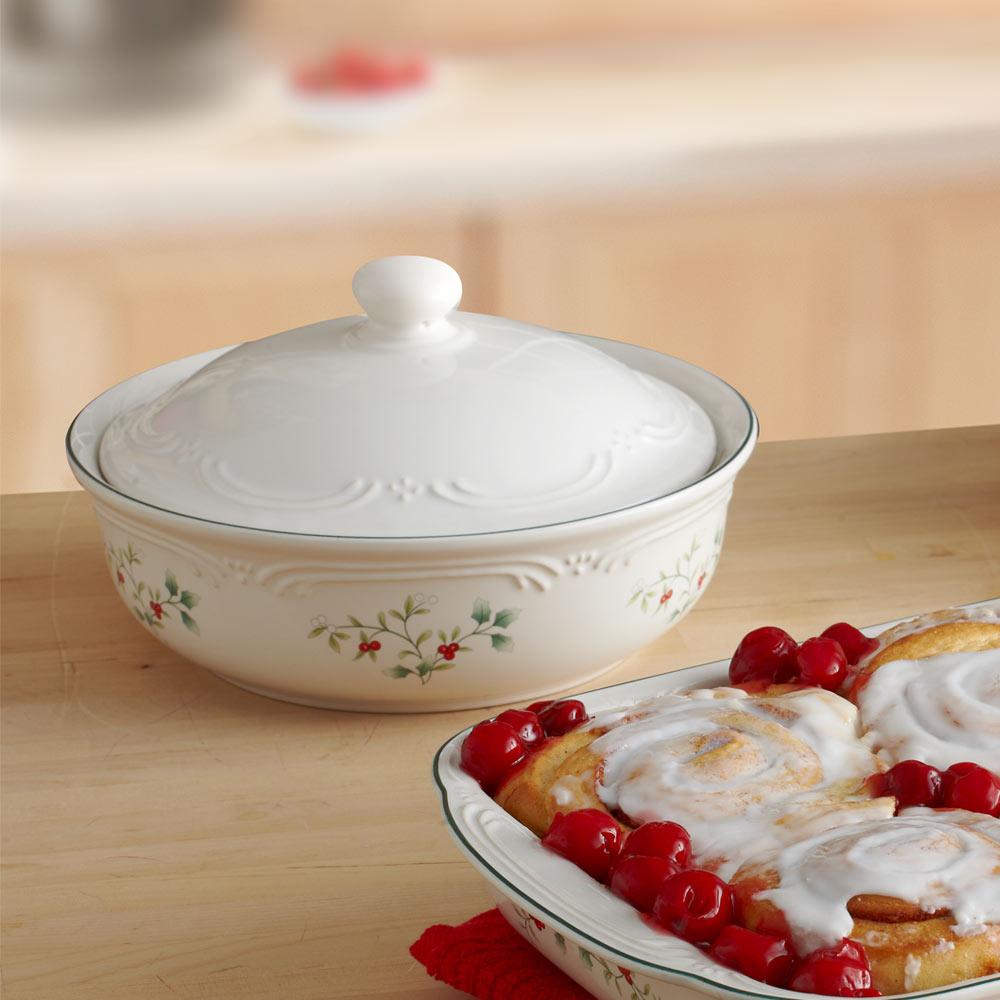 Winterberry round covered casserole in white porcelain with an embossed pattern, lid in place beside a plate of icing glazed rolls and cranberries.